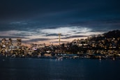 A nighttime cityscape featuring a waterfront with buildings illuminated by lights. The Space Needle is prominent, rising above the city. The sky is dark with lingering sunset hues.