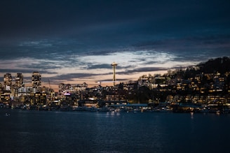 A nighttime cityscape featuring a waterfront with buildings illuminated by lights. The Space Needle is prominent, rising above the city. The sky is dark with lingering sunset hues.