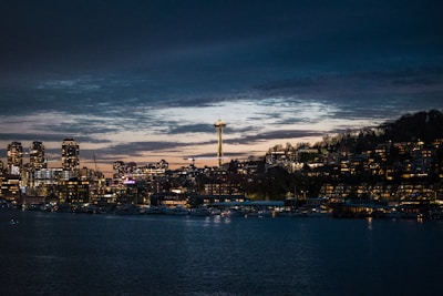 A nighttime cityscape featuring a waterfront with buildings illuminated by lights. The Space Needle is prominent, rising above the city. The sky is dark with lingering sunset hues.