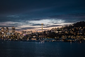 A nighttime cityscape featuring a waterfront with buildings illuminated by lights. The Space Needle is prominent, rising above the city. The sky is dark with lingering sunset hues.