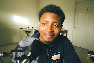 A person is sitting in front of a microphone with a slight smile, wearing a dark hoodie. The room is well-lit with natural light coming from a window, and there is a desk and chair in the background against a white wall.