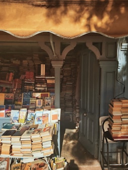 A cozy, small bookshop filled with towering stacks of books organized both on tables and inside open shelves. The scene is bathed in warm, golden light, with a partially open wooden door revealing more books stacked inside. The shop has an inviting, slightly cluttered but charming atmosphere.