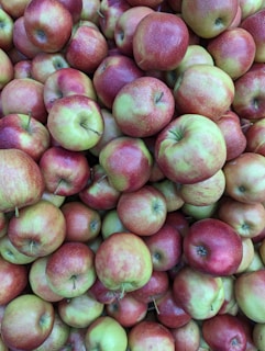 A large pile of freshly picked apples, predominantly red with hints of green. The apples have a glossy, smooth texture and are closely packed together, creating a vibrant display.
