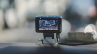 A dashboard-mounted camera capturing street footage, with a display showing a city street scene. The device is connected with a cable and positioned on a car dashboard against a blurred urban background.