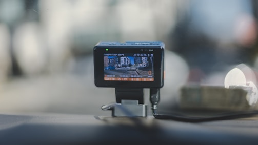 A dashboard-mounted camera capturing street footage, with a display showing a city street scene. The device is connected with a cable and positioned on a car dashboard against a blurred urban background.