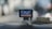 A dashboard-mounted camera capturing street footage, with a display showing a city street scene. The device is connected with a cable and positioned on a car dashboard against a blurred urban background.