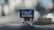 A dashboard-mounted camera capturing street footage, with a display showing a city street scene. The device is connected with a cable and positioned on a car dashboard against a blurred urban background.