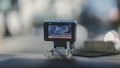 A dashboard-mounted camera capturing street footage, with a display showing a city street scene. The device is connected with a cable and positioned on a car dashboard against a blurred urban background.