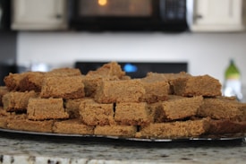 A stack of square-shaped baked goods, likely pieces of gingerbread or a similar treat, is arranged on a kitchen counter. The background features a microwave and part of a stove, suggesting a home kitchen setting.