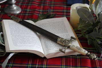 An open book rests on a red plaid fabric, with an ornate dagger placed across its pages. The dagger features intricate detailing on its handle and sheath. Surrounding the book are green leaves and a lit candle, creating a cozy ambience.