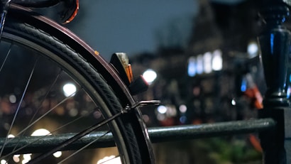 A close-up of a bicycle tire with intricate tread patterns, attached to a metal frame. The background features blurred city lights and a darkened urban environment, suggesting a nighttime setting.