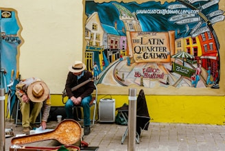 A colorful mural depicts a lively street scene in Galway's Latin Quarter, showcasing vibrant buildings and people. Two street musicians sit against the wall, one playing an instrument and the other interacting with items on the ground. Autumnal hues and lively urban elements dominate the scene.