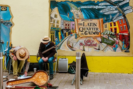 A colorful mural depicts a lively street scene in Galway's Latin Quarter, showcasing vibrant buildings and people. Two street musicians sit against the wall, one playing an instrument and the other interacting with items on the ground. Autumnal hues and lively urban elements dominate the scene.