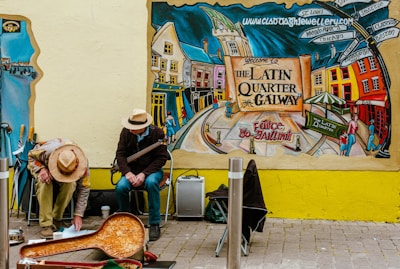 A colorful mural depicts a lively street scene in Galway's Latin Quarter, showcasing vibrant buildings and people. Two street musicians sit against the wall, one playing an instrument and the other interacting with items on the ground. Autumnal hues and lively urban elements dominate the scene.