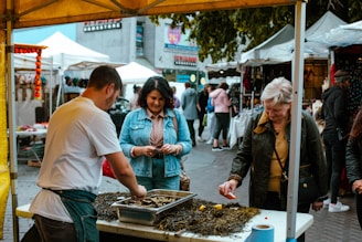 A bustling market scene with several people interacting around a food stall. A man is preparing seafood on a table covered with seaweed, while two women are engaged and observing the process. An array of tents and stalls in the background suggests a lively outdoor market environment.
