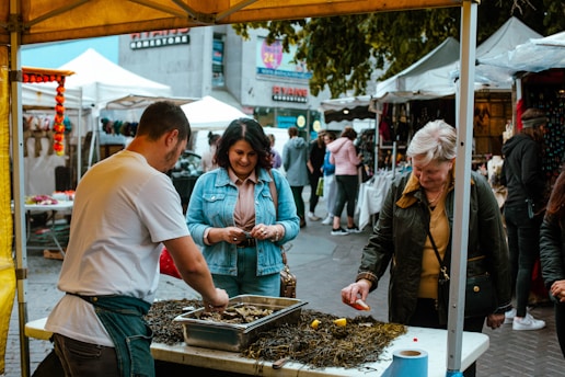 A bustling market scene with several people interacting around a food stall. A man is preparing seafood on a table covered with seaweed, while two women are engaged and observing the process. An array of tents and stalls in the background suggests a lively outdoor market environment.