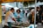 A bustling market scene with several people interacting around a food stall. A man is preparing seafood on a table covered with seaweed, while two women are engaged and observing the process. An array of tents and stalls in the background suggests a lively outdoor market environment.