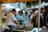 A bustling market scene with several people interacting around a food stall. A man is preparing seafood on a table covered with seaweed, while two women are engaged and observing the process. An array of tents and stalls in the background suggests a lively outdoor market environment.