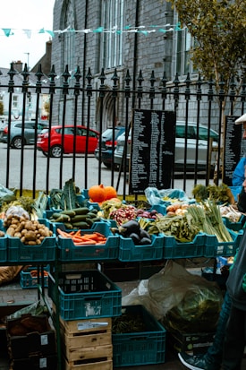 A variety of fresh vegetables are displayed in blue crates at an outdoor market setup. The assortment includes carrots, potatoes, eggplants, radishes, and pumpkins. The setting is near a stone building with iron railings and parked cars visible in the background. A chalkboard with a list is placed beside the vegetables.