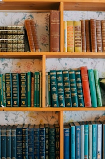 A wooden bookshelf holds several rows of hardcover books of varying colors, primarily green, blue, and brown. The spines have intricate designs and some display gold text. The bookshelf is set against a floral patterned wallpaper.