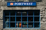 A store window with a prominent blue frame and stone facade displays various colorful clothing items inside. The signage above the window includes a blue 'PORTWEST' sign, and there is also a 'Columbia' logo visible above the clothing.