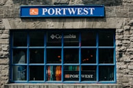 A store window with a prominent blue frame and stone facade displays various colorful clothing items inside. The signage above the window includes a blue 'PORTWEST' sign, and there is also a 'Columbia' logo visible above the clothing.