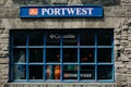 A store window with a prominent blue frame and stone facade displays various colorful clothing items inside. The signage above the window includes a blue 'PORTWEST' sign, and there is also a 'Columbia' logo visible above the clothing.