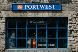 A store window with a prominent blue frame and stone facade displays various colorful clothing items inside. The signage above the window includes a blue 'PORTWEST' sign, and there is also a 'Columbia' logo visible above the clothing.