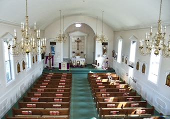 An interior view of a church featuring wooden pews arranged in rows on a green carpet. The altar area is adorned with religious icons and a crucifix. Several chandeliers with multiple candle-like bulbs hang from the arched ceiling. The walls are decorated with framed religious artwork, and light filters in through tall, narrow windows.