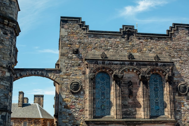 An ancient stone building with detailed architecture, featuring large arched windows with intricate designs and decorative elements. The structure is made of various shades of stone, with some carvings or sculptures visible on the facade. A blue sky provides a backdrop, adding contrast to the stone's texture.
