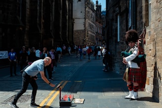 A street performer dressed in traditional Scottish attire, including a kilt and hat, plays the bagpipes on a cobblestone street. A man bends down to place money in a collection case. People are walking in the background, with historical stone buildings lining the street.