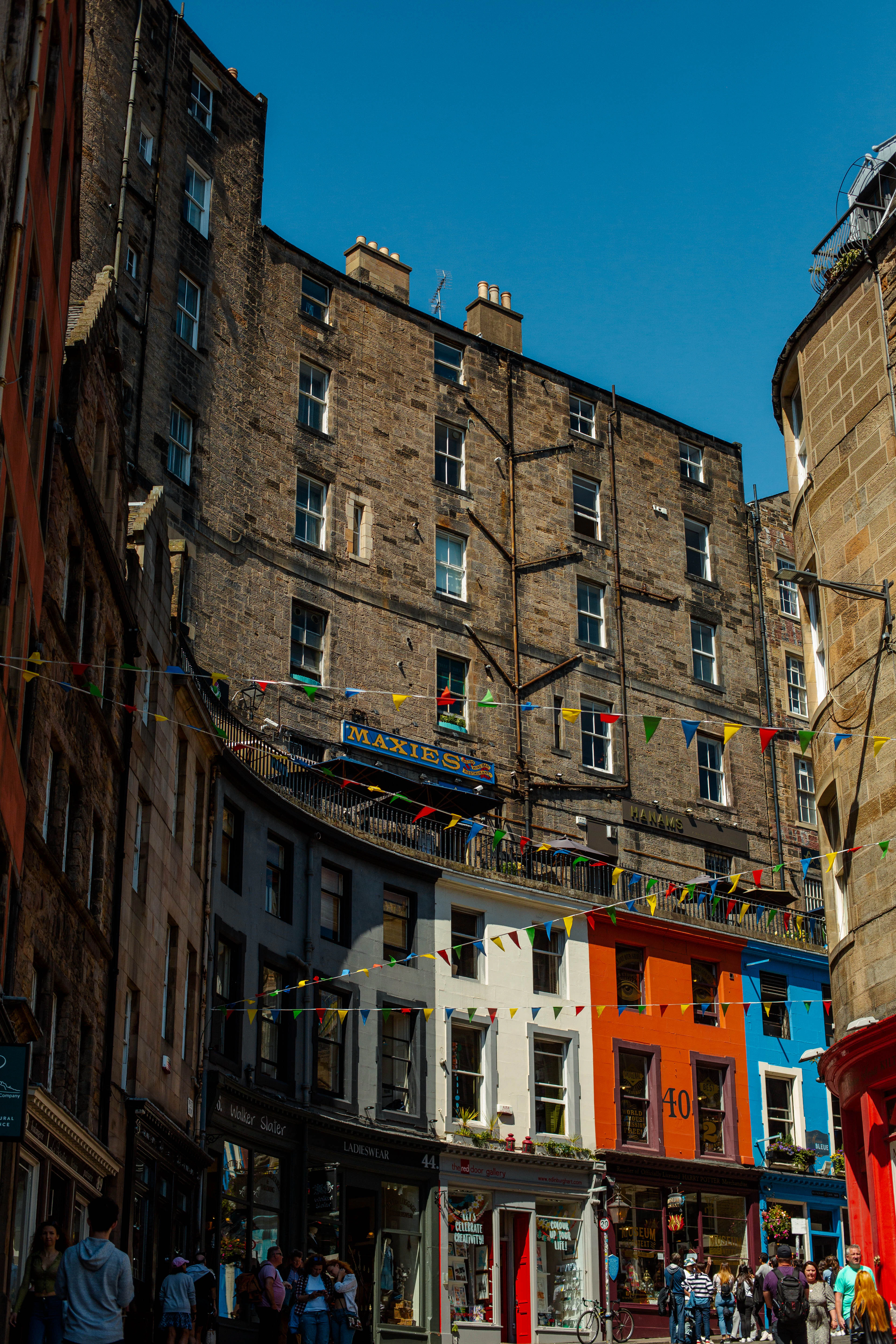 Bunting strings over a Grassmarket street of orange and blue tenement shopfronts.