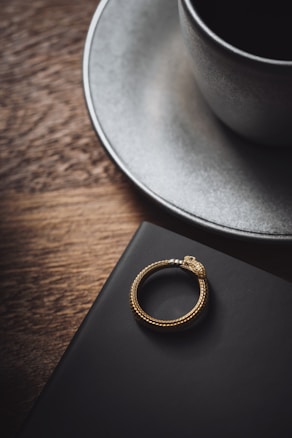A gold snake-shaped ring rests on a dark surface next to a ceramic cup on a saucer with a wooden background, conveying a sense of luxury and elegance.
