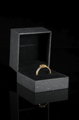 A gold ring with intricate details rests inside an open black velvet-lined jewelry box. The box is placed on a reflective surface, enhancing the elegance of the scene.