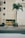 A tall, beige building with vertical lines designed into its facade. In front of the building, two yellow dumpsters are placed against a closed garage door. A lone palm tree stands on the right side near some shrubs on the ground. The scene is set along a deserted street with a sidewalk leading up to the building.