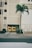 A tall, beige building with vertical lines designed into its facade. In front of the building, two yellow dumpsters are placed against a closed garage door. A lone palm tree stands on the right side near some shrubs on the ground. The scene is set along a deserted street with a sidewalk leading up to the building.