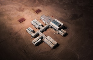 Aerial view of a structure composed of connected modular units resembling shipping containers, arranged in a cross-like formation on a barren, sandy landscape. A small red flag is visible on top of the central unit.