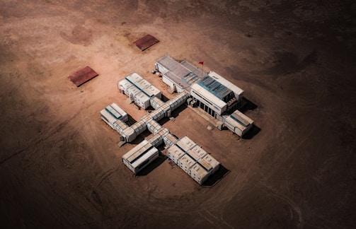 Aerial view of a structure composed of connected modular units resembling shipping containers, arranged in a cross-like formation on a barren, sandy landscape. A small red flag is visible on top of the central unit.