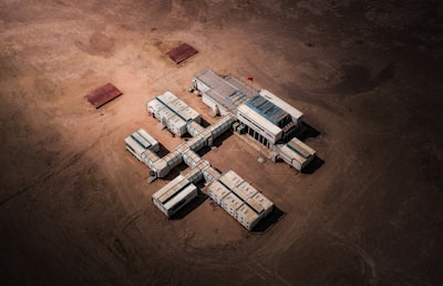 Aerial view of a structure composed of connected modular units resembling shipping containers, arranged in a cross-like formation on a barren, sandy landscape. A small red flag is visible on top of the central unit.