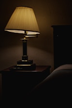 A dimly lit table lamp with a pleated shade sits on a wooden nightstand, casting a soft warm glow. The surrounding area is shadowed, with part of a bed visible on the right.