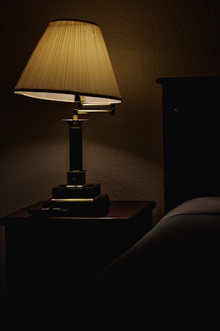 A dimly lit table lamp with a pleated shade sits on a wooden nightstand, casting a soft warm glow. The surrounding area is shadowed, with part of a bed visible on the right.