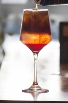 A glass is filled with a colorful layered drink, consisting of shades of red and brown, being poured from a can. The light in the background creates a soft glow, highlighting the condensation and swirling patterns within the drink.