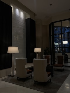 A modern, dimly lit lounge area featuring a cluster of high-backed chairs arranged around small tables. Tall, elegant floor lamps provide soft lighting, while dark and light wall panels create a stylish contrast. A large window shows a glimpse of the building's exterior.