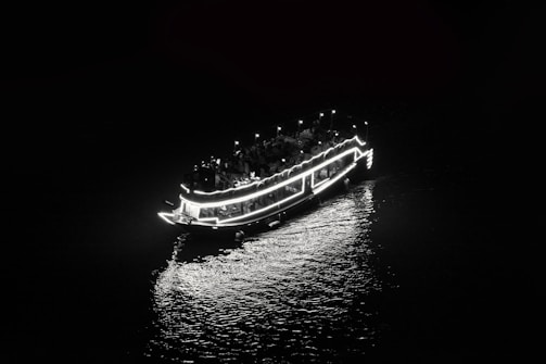 A boat adorned with bright lights is cruising at night, creating a luminous reflection on the water below. The scene is captured in a monochrome setting, emphasizing the contrast between light and dark.
