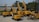A collection of yellow construction excavators and heavy machinery is parked on a concrete surface. The machines are branded with 'SANY' and are aligned in rows with some positioned at different angles. In the background, there are trees and a partially visible building.