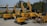A collection of yellow construction excavators and heavy machinery is parked on a concrete surface. The machines are branded with 'SANY' and are aligned in rows with some positioned at different angles. In the background, there are trees and a partially visible building.