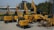 A collection of yellow construction excavators and heavy machinery is parked on a concrete surface. The machines are branded with 'SANY' and are aligned in rows with some positioned at different angles. In the background, there are trees and a partially visible building.
