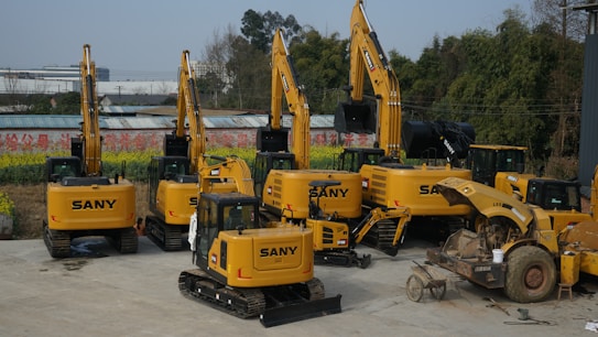 A collection of yellow construction excavators and heavy machinery is parked on a concrete surface. The machines are branded with 'SANY' and are aligned in rows with some positioned at different angles. In the background, there are trees and a partially visible building.