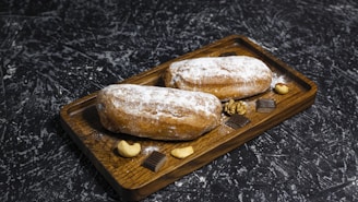 Two bread rolls dusted with powdered sugar are placed on a wooden tray, surrounded by assorted nuts and pieces of chocolate. The background is a textured dark surface.