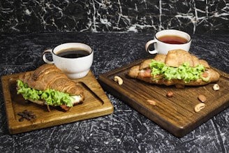 Two wooden boards each hold a croissant stuffed with fresh lettuce and slices of turkey or ham. One board accompanies a cup filled with black coffee, and the other features a cup of tea. Surrounding the boards, there are scattered nuts and coffee beans, all set against a dark marbled background.